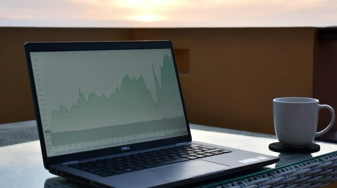 A Dell laptop on an outdoor table showing a green area chart, a white mug beside it, low sun over the water.