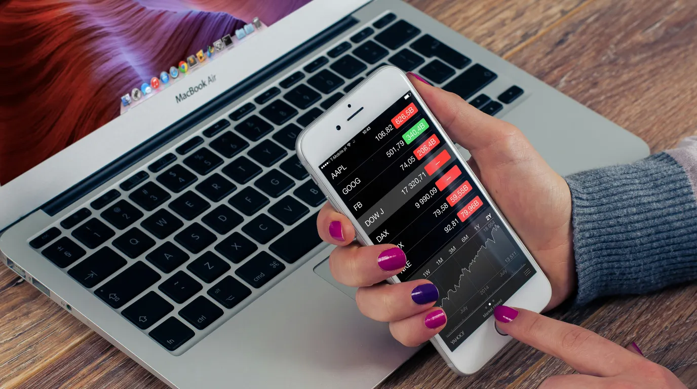 A manicured hand holds a smartphone listing share tickers in red and green above an open laptop keyboard on a wooden desk.