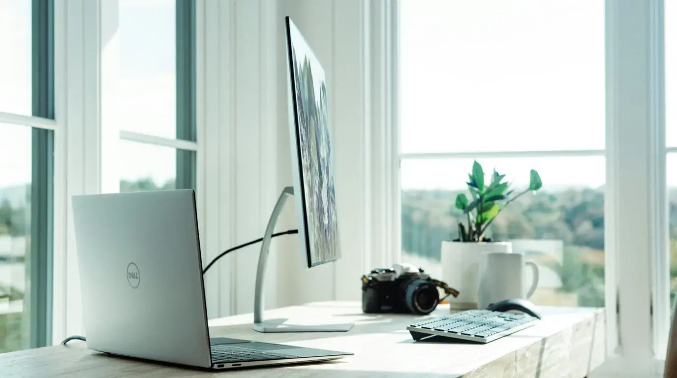 A pale desk by a window with an open laptop, a camera and a mug