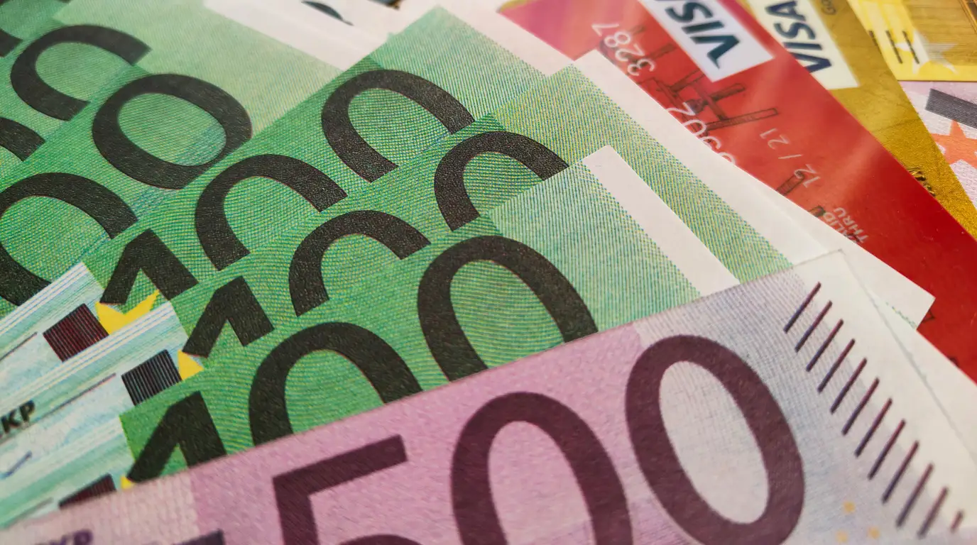 Close-up of green hundred-euro notes fanned over a lilac five-hundred note, with payment cards showing at the top edge.