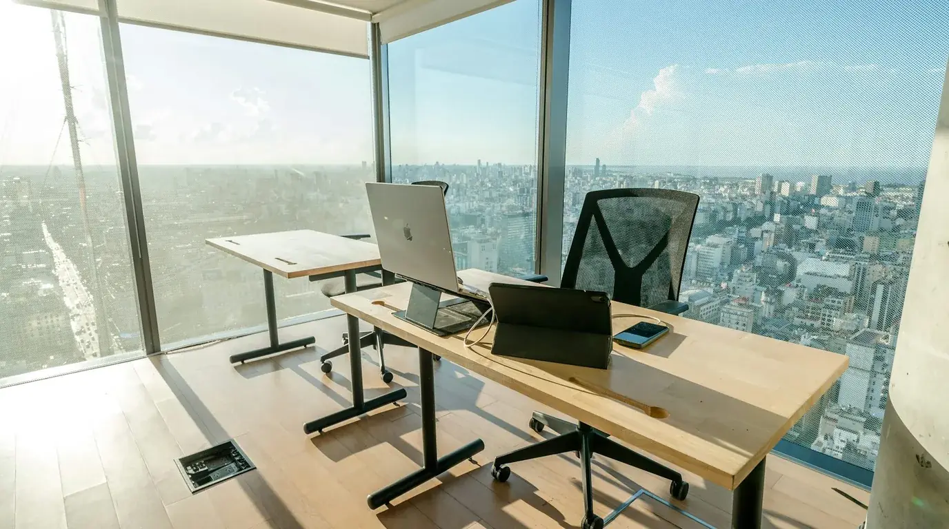 A bright corner office with a laptop on a pale desk and a city view through full-height glass