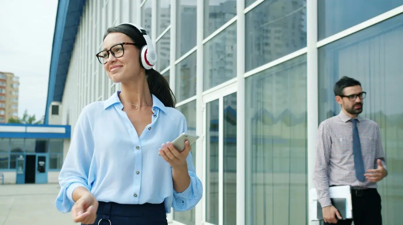 A woman in headphones walking past a glass office facade with her phone