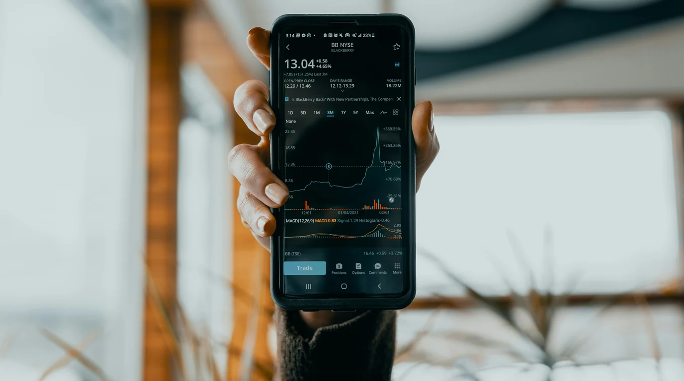 A hand holds up a phone showing a dark trading screen with a spiky price chart and a blue trade button, the room blurred behind.