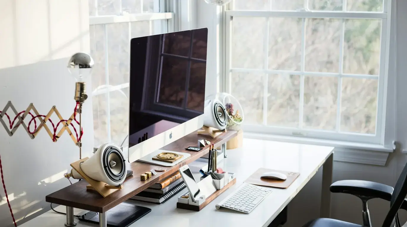 A bright desk by a window with a monitor, speakers and a keyboard