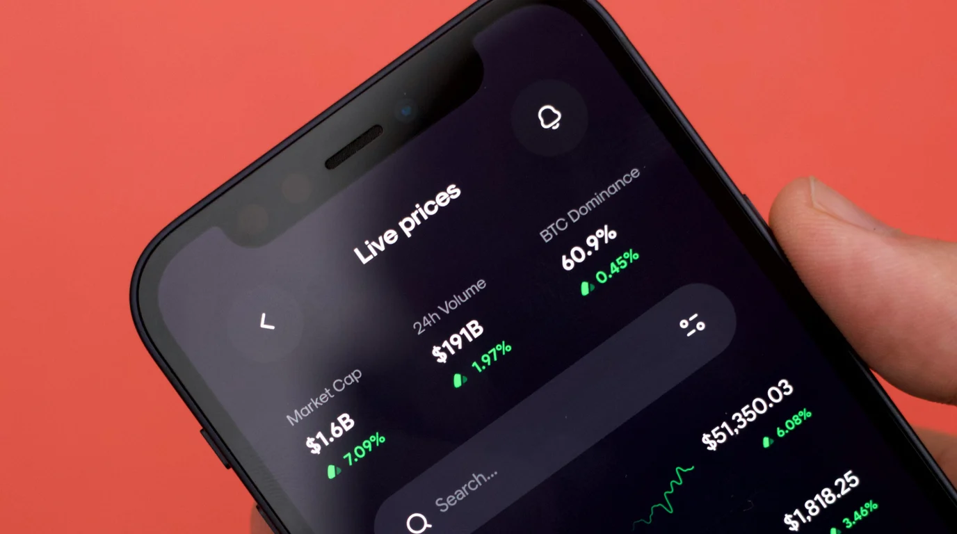 A hand tilts a phone against a coral background, its dark screen listing live prices, market cap and volume in white and green.