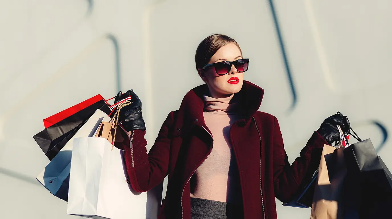 A woman in a burgundy coat and sunglasses holding several paper shopping bags in each hand
