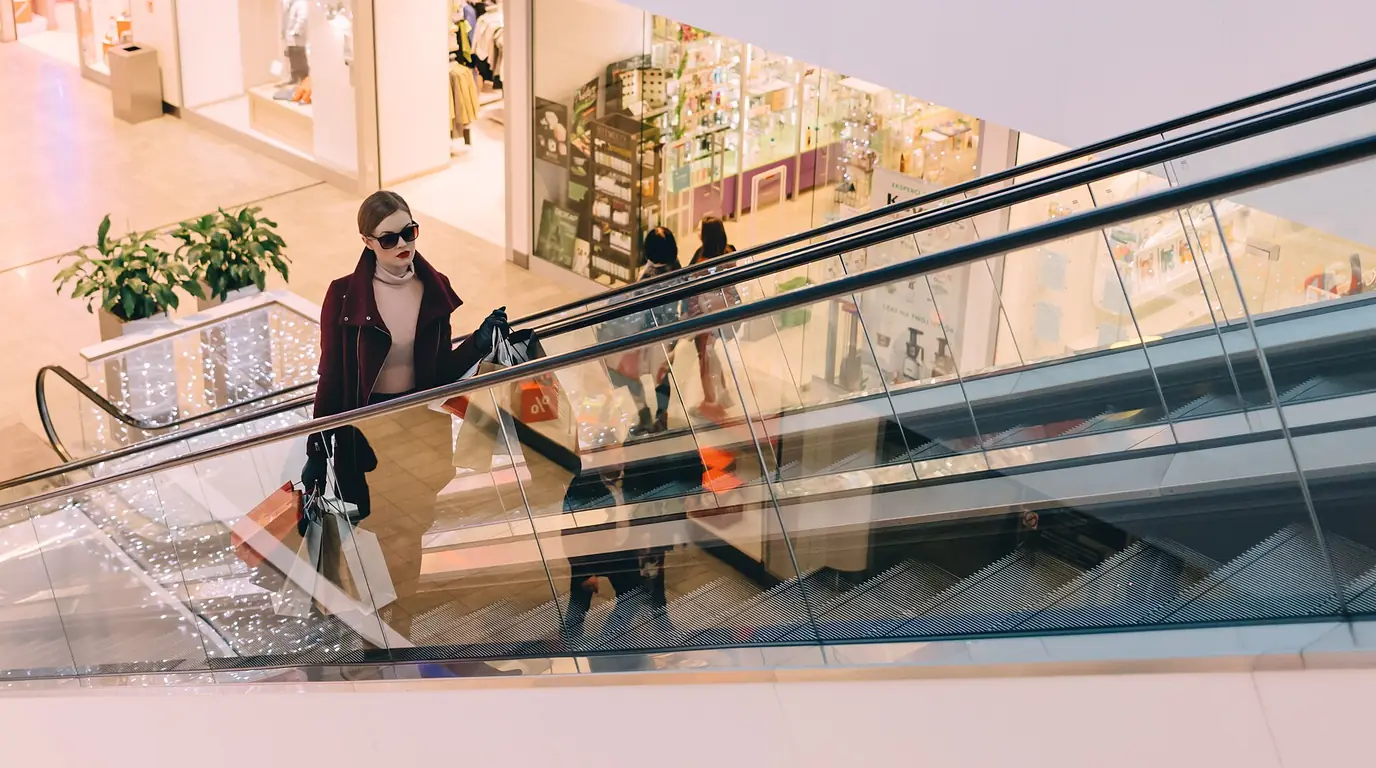 A shopper carrying several bags rides an escalator in a bright shopping mall.