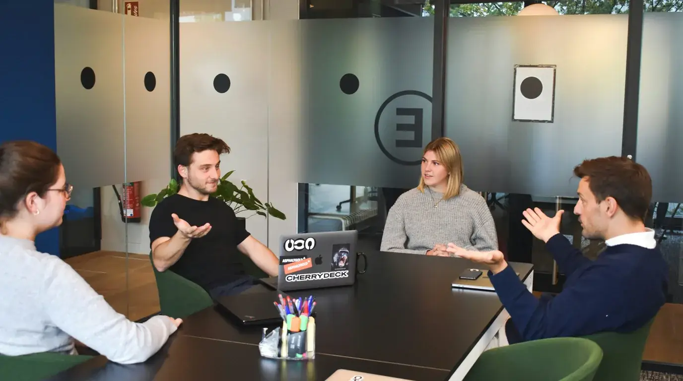 Four people in discussion around a small table in a glass-walled room