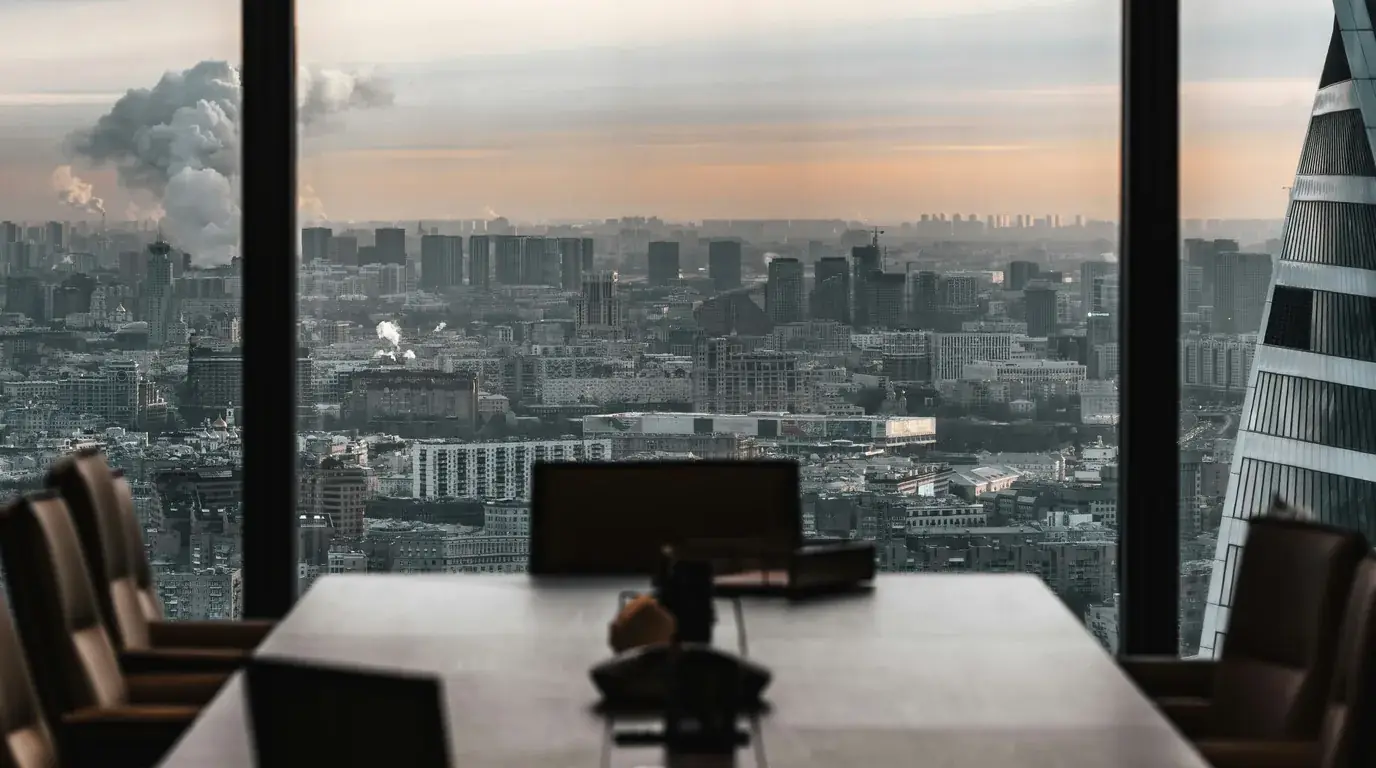 An empty meeting room high in a tower, a city stretching away beyond the glass under a low warm sky.