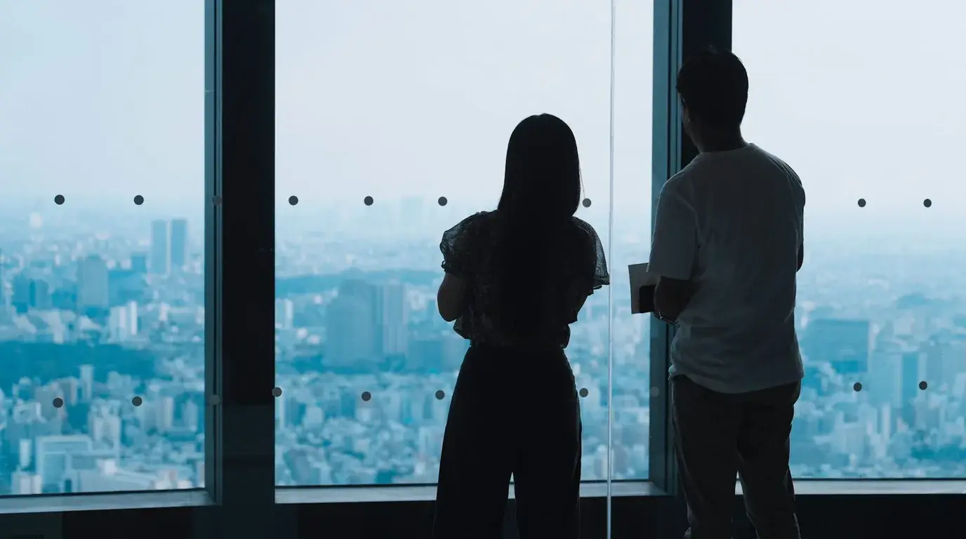 Two people silhouetted at a floor-to-ceiling window, looking out over a hazy low-rise city from high up.