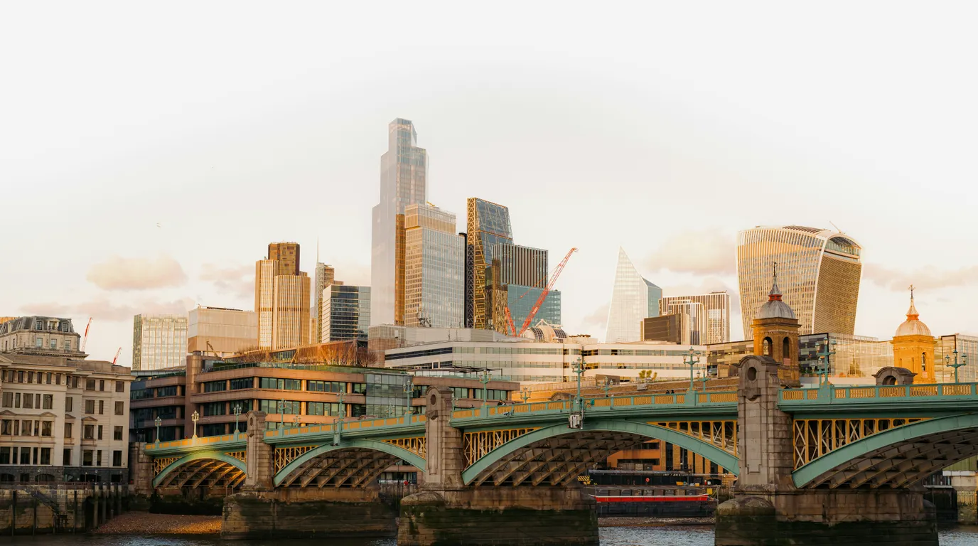 Green ironwork arches of a river bridge in low sun, with a skyline of glass towers and a domed church behind.